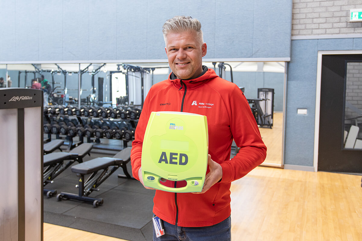 Sportinstructeur van het Alfa college poseert met een AED in de gym